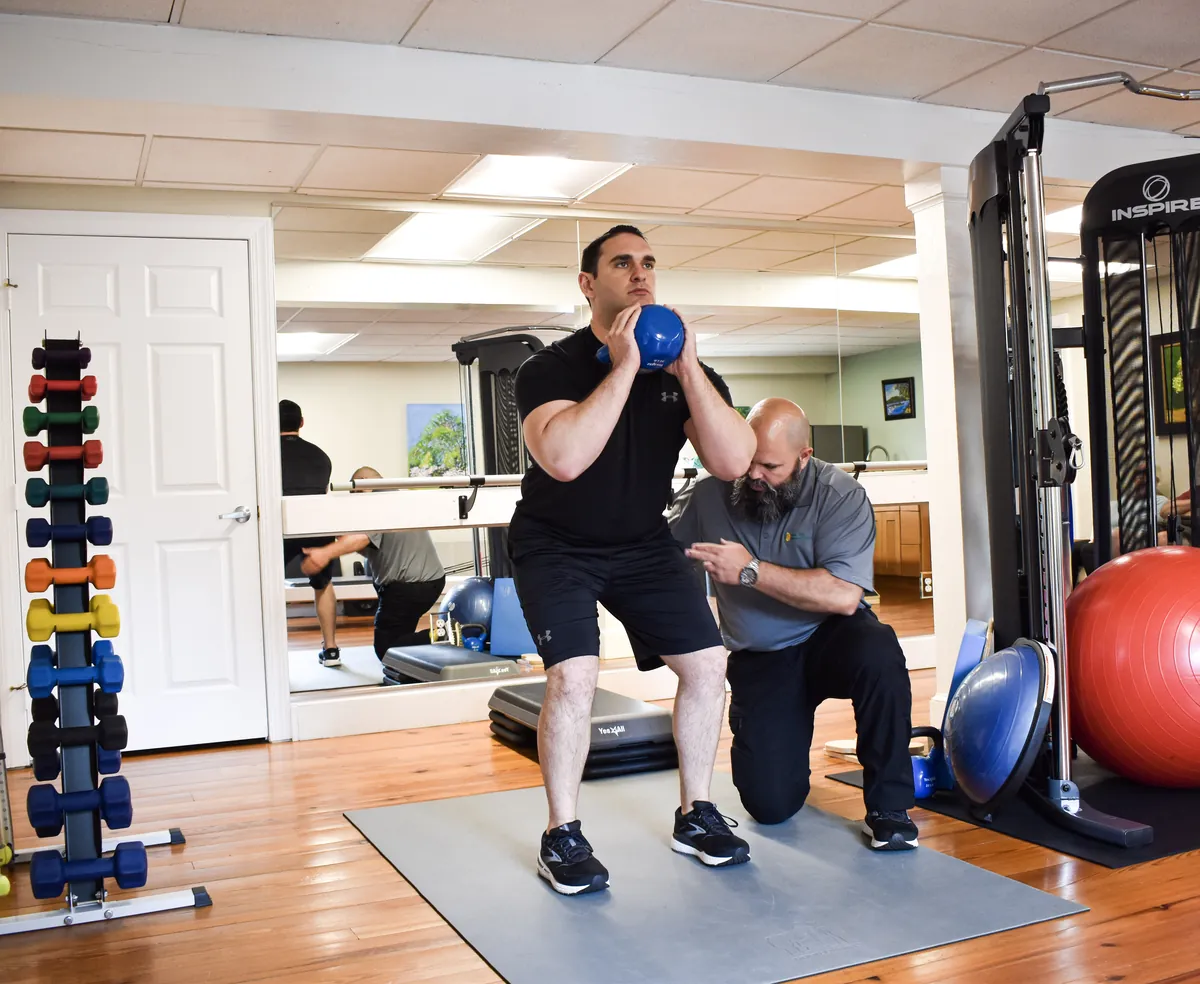Physical therapist coaching a client through a weighted squat during a maintenance program session